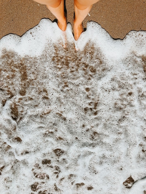 Woman legs barefoot at sea foam waves on sand beach summer day. top view above women feet