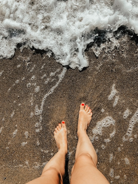 woman legs barefoot at sea foam waves on sand beach summer day. top view above women feet