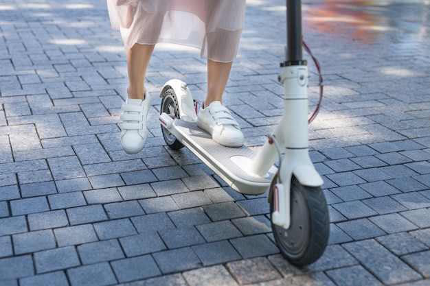 Woman legs on eco friendly electro scooter on sidewalk tile in\
sunny day