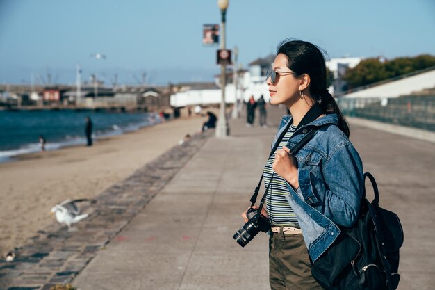 woman lens man holding camera smiling cheerfully sightseeing in port in pier 39 san francisco. young asian girl backpacker joyful looking enjoy the ocean sea view. wild freedom animal seagull resting