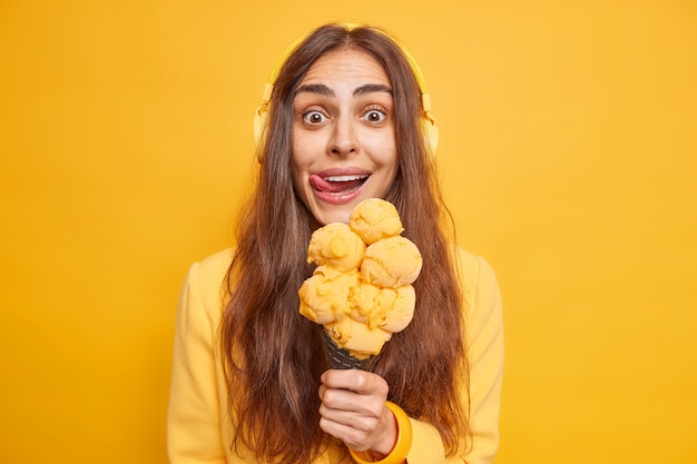 Woman licks lips with tongue feels temptation to eat delicious\
ice cream in waffle enjoys summer dessert during walk listens music\
isolatd on yellow