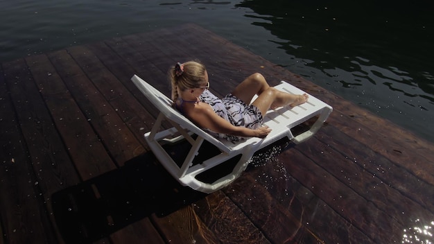 Woman lie on a sunbed in sunglasses and a boho silk shawl Girl rest on a flood wood underwater pier The pavement is covered with water in the lake In the background are mountain and a forest