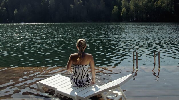 Photo woman lie on a sunbed in sunglasses and a boho silk shawl girl rest on a flood wood underwater pier the pavement is covered with water in the lake