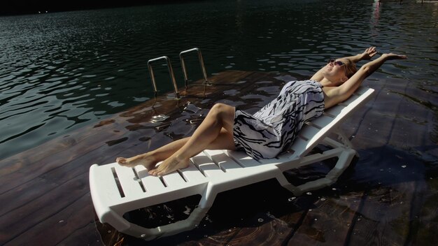 Woman lie on a sunbed in sunglasses and a boho silk shawl Girl rest on a flood wood underwater pier The pavement is covered with water in the lake