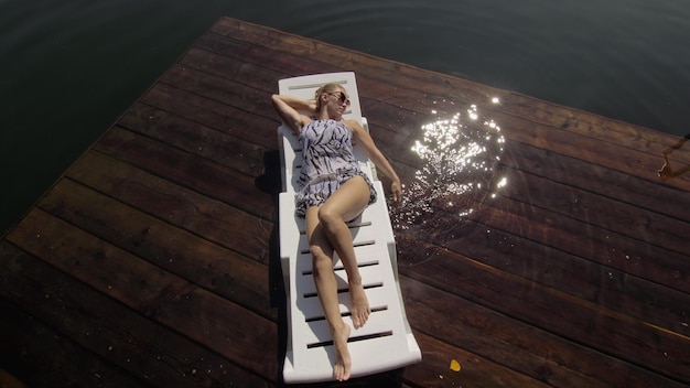 Woman lie on a sunbed in sunglasses and a boho silk shawl Girl rest on a flood wood underwater pier The pavement is covered with water in the lake