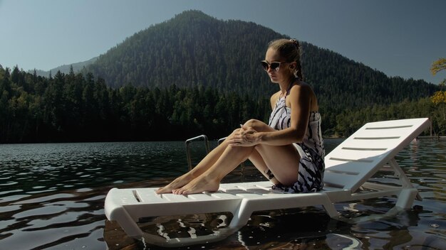 Woman lie on a sunbed in sunglasses and a boho silk shawl Girl rest on a flood wood underwater pier The pavement is covered with water in the lake