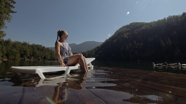 Woman lie on a sunbed in sunglasses and a boho silk shawl Girl rest on a flood wood underwater pier The pavement is covered with water in the lake