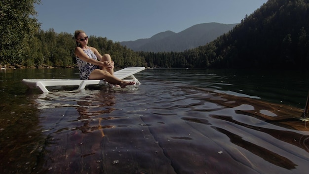 Женщина лежит на шезлонге в солнцезащитных очках и шелковой шали в стиле бохо Девушка отдыхает на подводном пирсе из затопленного дерева Тротуар в озере покрыт водой