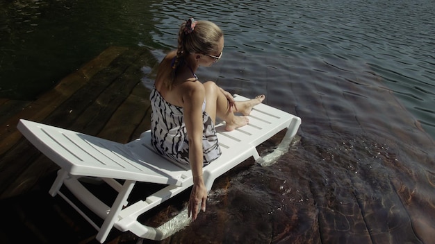 Woman lie on a sunbed in sunglasses and a boho silk shawl Girl rest on a flood wood underwater pier The pavement is covered with water in the lake