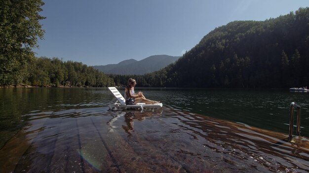 Woman lie on a sunbed in sunglasses and a boho silk shawl Girl rest on a flood wood underwater pier The pavement is covered with water in the lake