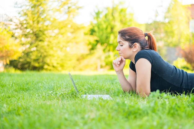 公園でうつぶせになり、ラップトップでタイプする女性 フリーランサーの女性が屋外でリモートで働く 芝生の上でコンピューターで勉強する少女