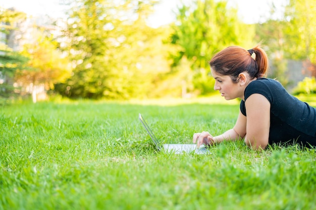 A woman lies on her stomach in a park and types on a laptop Female freelancer works remotely outdoors Girl is studying on a computer on the lawn