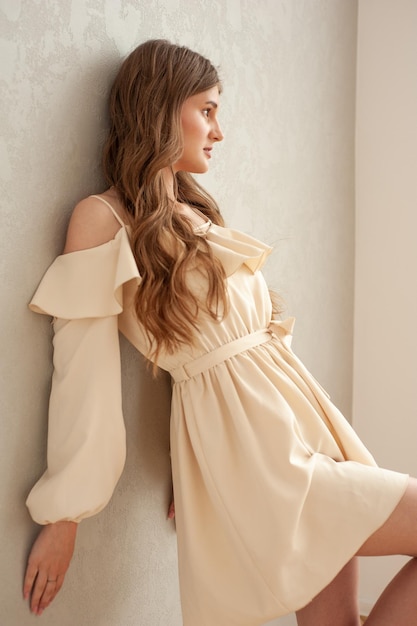 A woman in a light beige dress leans against a wall.