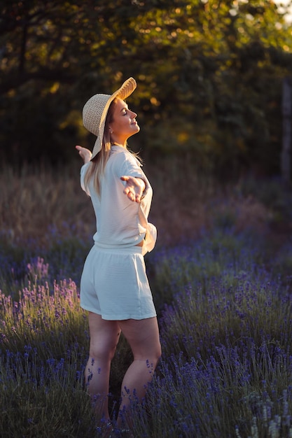 가벼운 여름 흰색 양복과 모자를 쓴 여성이 라벤더 밭에서 석양을 만나기 위해 손을 펼쳤습니다.