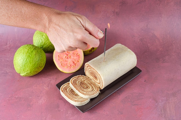 Woman lighting a candle sparkles on a cake roll next to guavas (top view).