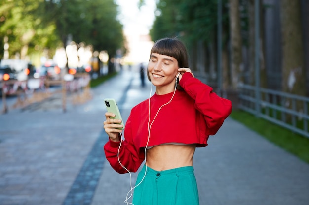 woman listening music on the street with phone, cute millenial woman in red stylish sweater having smartphone and earphone and listen music