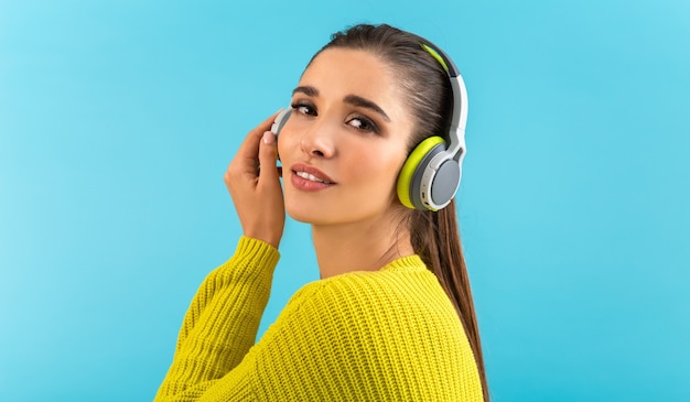 woman listening to music in wireless headphones happy wearing yellow knitted sweater posing on blue