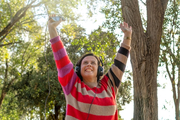 Photo woman listening to music with headphones in the park raising an arm concept of happiness vitality