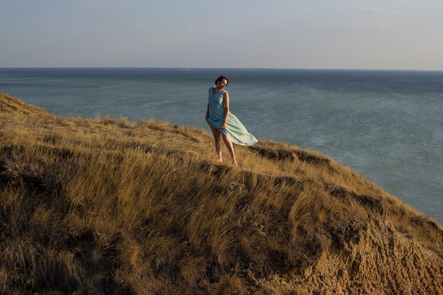 woman in a long blue dress standing on a high cliff by the sea or ocean on sunny windy summer day.