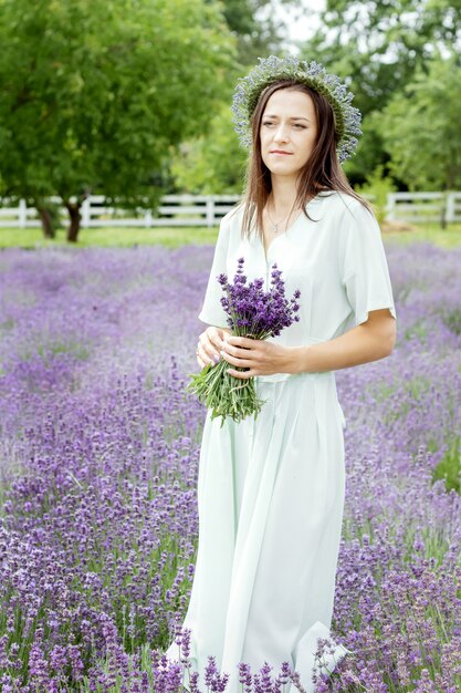 長いオリーブのドレスとラベンダー畑の帽子の女性花輪とラベンダーの花束を持つ若い女性