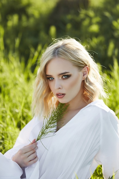 woman in a long white dress sits on the grass in a field. Blonde woman in the sun in a light dress. woman resting and dreaming, perfect summer makeup on her face