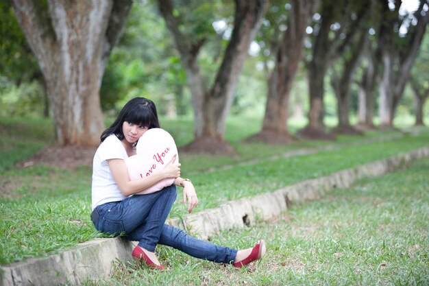 写真 公園の木に寄りかかって座っているあなたに愛を込めてクッションを握っている女性