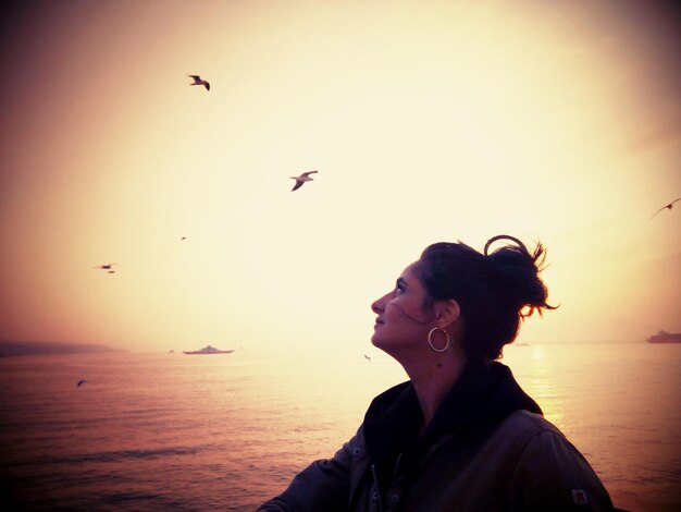 Photo woman looking at birds flying over sea against sky during sunset