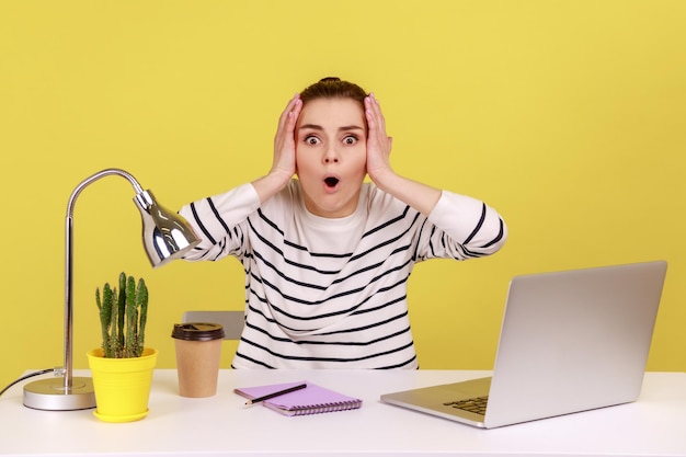 Woman looking at camera with big eyes and open mouth keeps hands on head problems with her work