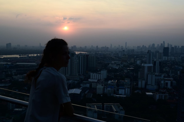 Woman looking at city buildings against sky during sunset