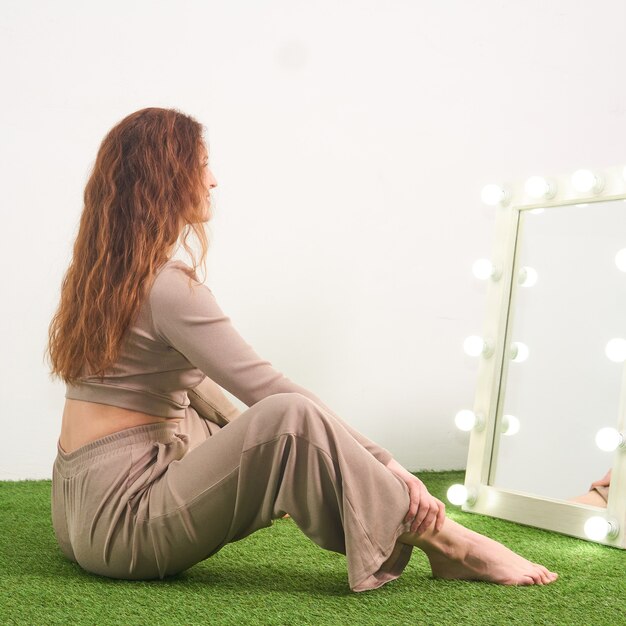 Woman looking at her reflection in illuminated mirror for dressing room sitting on floor in studio