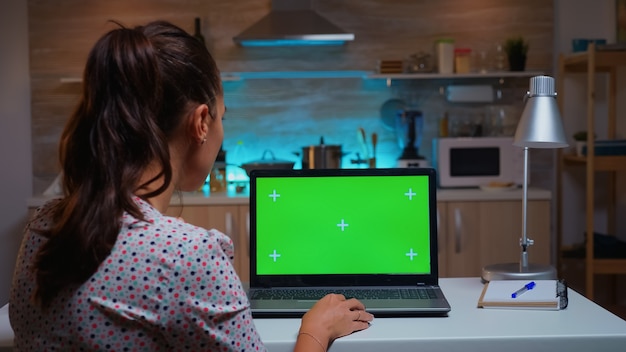 Woman looking at laptop with green mockup during night time in home kitchen. Freelancer watching desktop monitor display with green screen, chroma key, during night time working overtime.