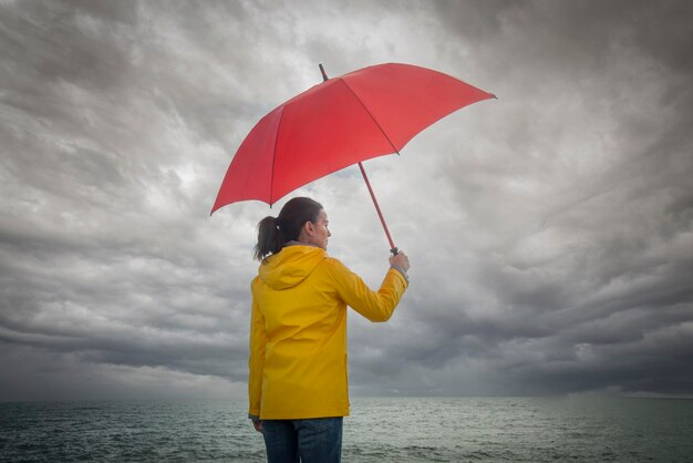 Photo woman looking out to see and a stormy sky holding a red umbrella and wearing a yellow coat