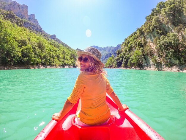 Woman looking over shoulder sitting on red canoe exploring calm tropical bay against backdrop of mountains valley. Woman sitting on red canoe on lake admiring scenic nature and mountain