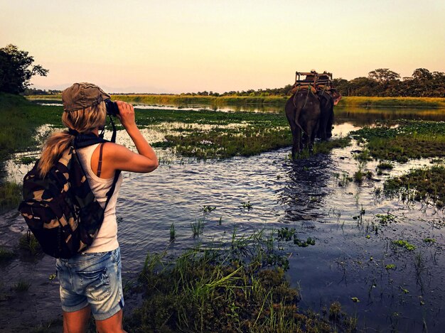 Photo woman looking through binoculars at lakeshore while elephant walking in background