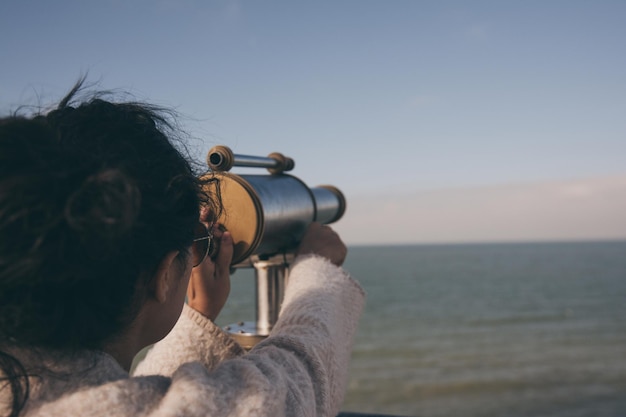 Woman looking through coin-operated binoculars at sea against sky