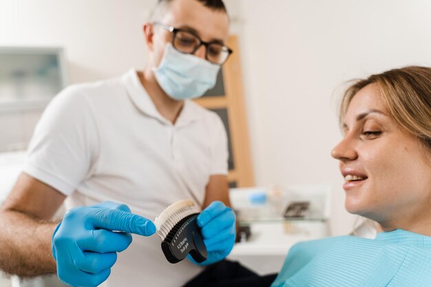 ベニヤを見ている女性、または医師の手で歯のカラーマッチングサンプルを移植している女性歯科歯科医は、歯科医院の女性患者のための歯のホワイトニングのための歯のカラーシェードガイドを示しています