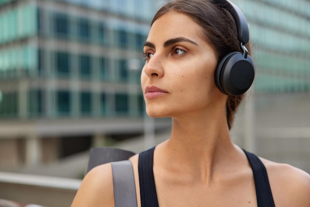 Woman looks away has pensive expression walks outdoors carries\
fitness mat prepares for workout poses outdoors outdoor listens\
music