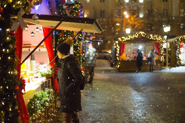 A woman looks and buys at a kiosk, people walk at the Christmas market in the festive decor of the streets. Exhibition and shopping malls in the city center. New Year.