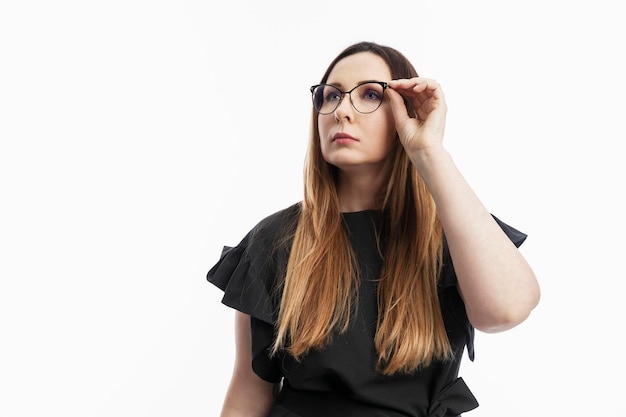 The woman looks up and adjusts her glasses. Serious brunette in a black blouse. Mindfulness in doing business. White background.