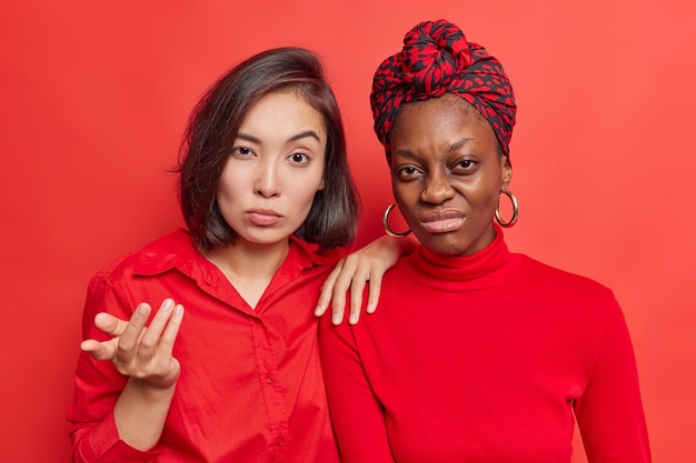  woman looks with puzzlement gestures indignant near female friend. Dissatisfied African American model doesnt like bad news frowns face poses on vivid red 