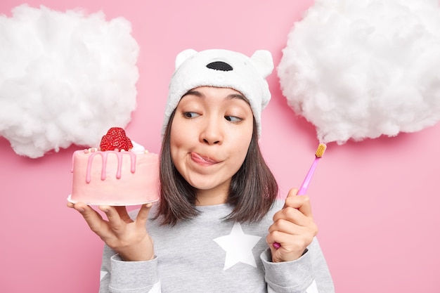 Woman looks with temptation at delicious strawberry cake licks\
lips holds toothbrush wants to have healthy teeth