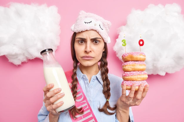 woman looks with worried expression celebrates 30th anniversary holds pile of donuts and milk wears sleepmask birthday ribbon isolated on pink