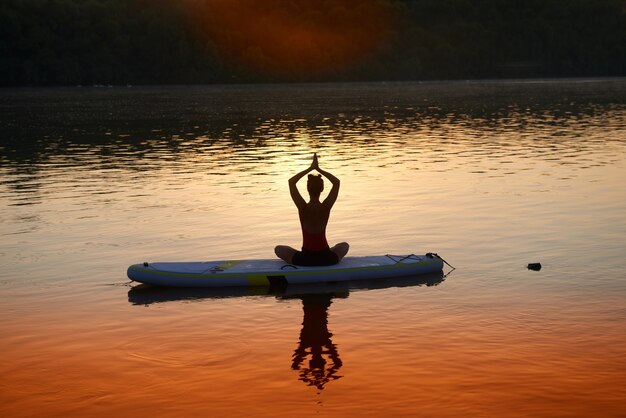 夕日を背景に瞑想するパデルボードに座っている蓮華座の女性