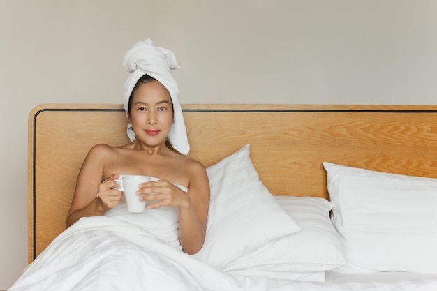 Photo woman lying in bed holding coffee mug with a towel on her head