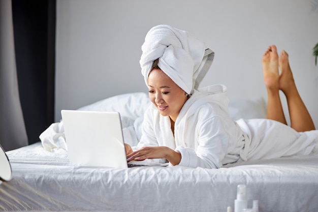 Woman lying on bed at home and working on her laptop computer, wearing towel and bathrobe, in the morning. working from home, quarantine coronavirus concept