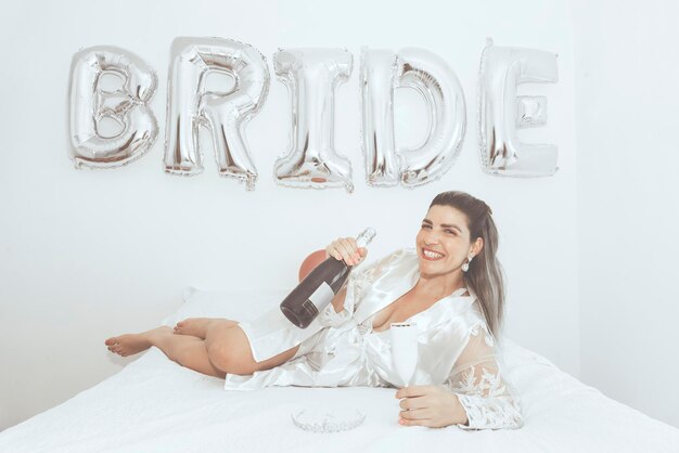 Photo woman lying in bed with glass of wine in hand and bottle of drink against white background written bride salvador bahia brazil                               person