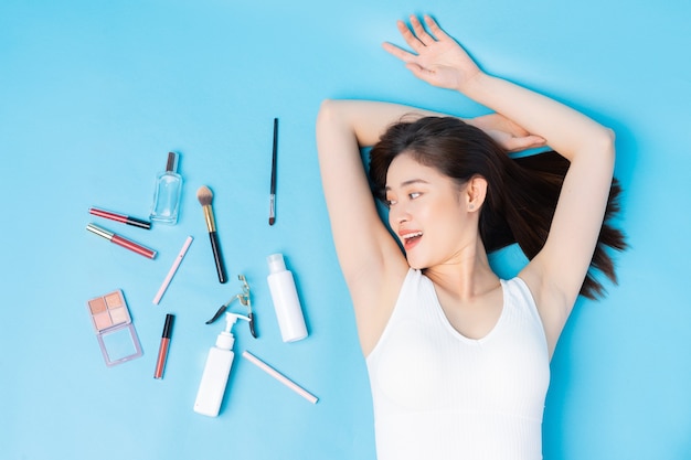 Woman lying on blue with skin care tools and makeup beside