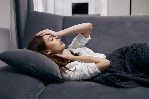 Женщина, лежащая дома на диване, покрытая одеялом, чувствует себя нездоровой рукой на лице