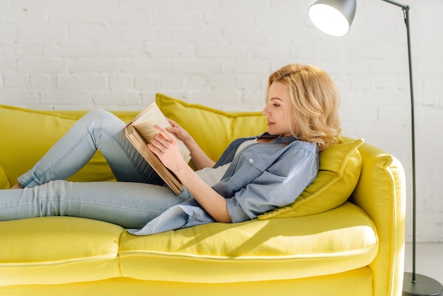 Фото Женщина лежит на уютном желтом диване и читает книгу