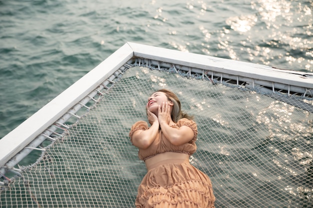 바다 배경으로 부두에 누워 있는 여자는 바다 옆 그물에 앉아 있습니다.